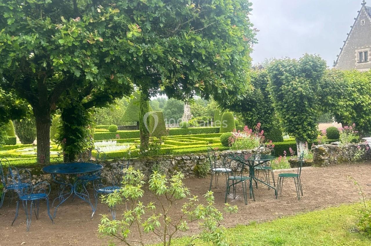 Jardin avec tables et chaises en métal bleu sous un arbre, entouré de haies taillées et de parterres fleuris.