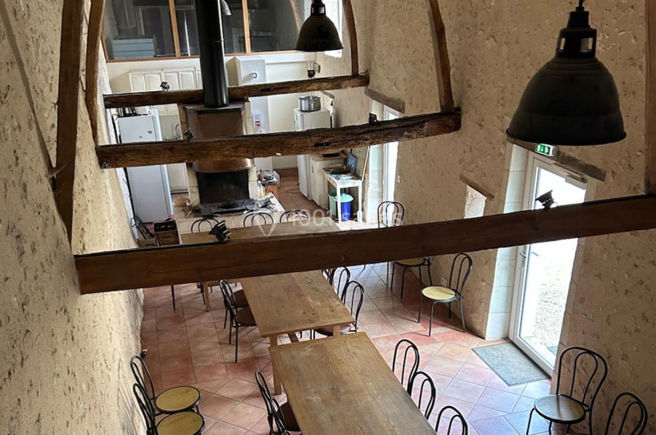 Salle à manger rustique avec poutres apparentes, tables en bois, chaises en métal et cuisine ouverte en arrière-plan.