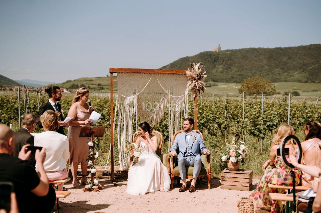 Un couple assis lors d'une cérémonie de mariage en plein air, devant une arche décorée, entouré d'invités.