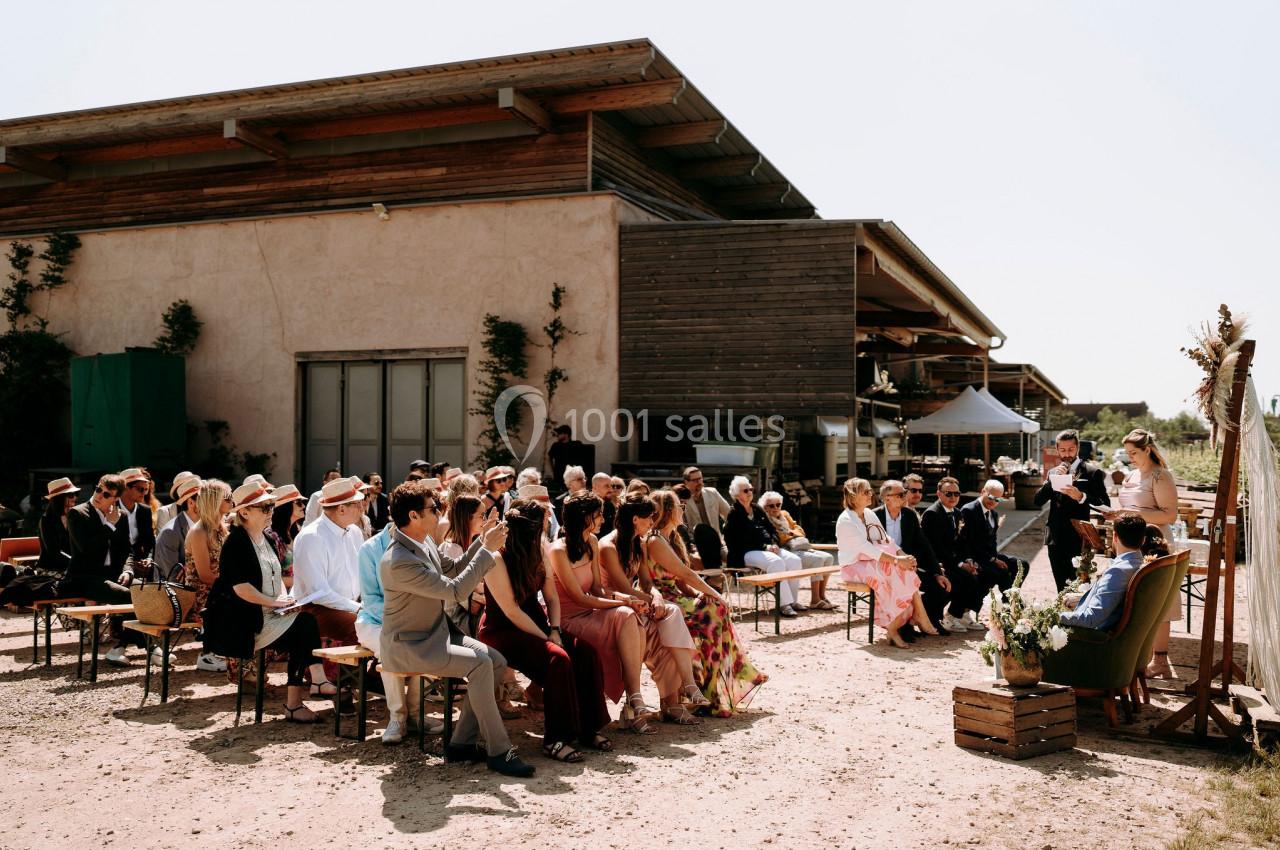 Cérémonie en plein air avec des invités assis, face à un couple et un officiant devant un bâtiment en bois.