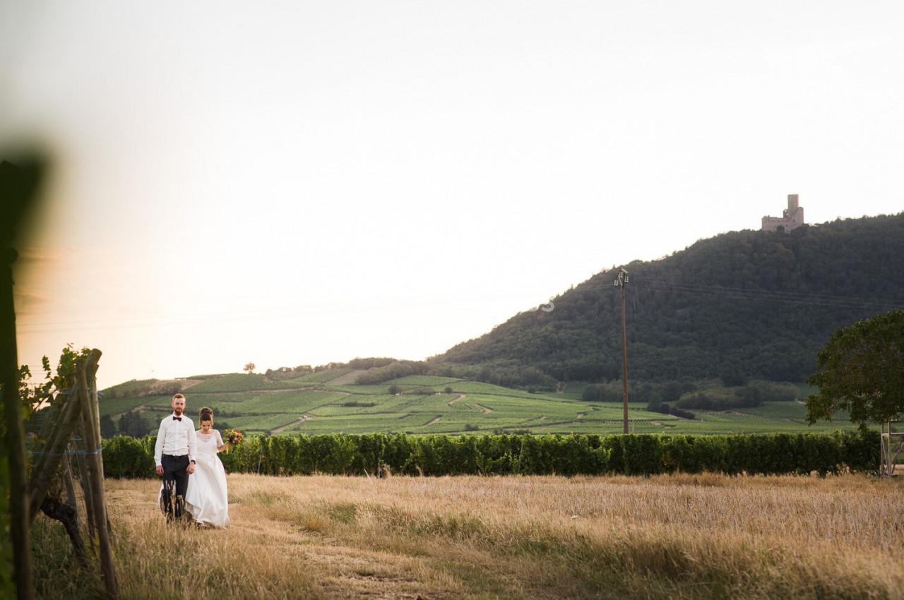 Un couple marche dans un champ près de vignes, avec une colline et un château en arrière-plan.