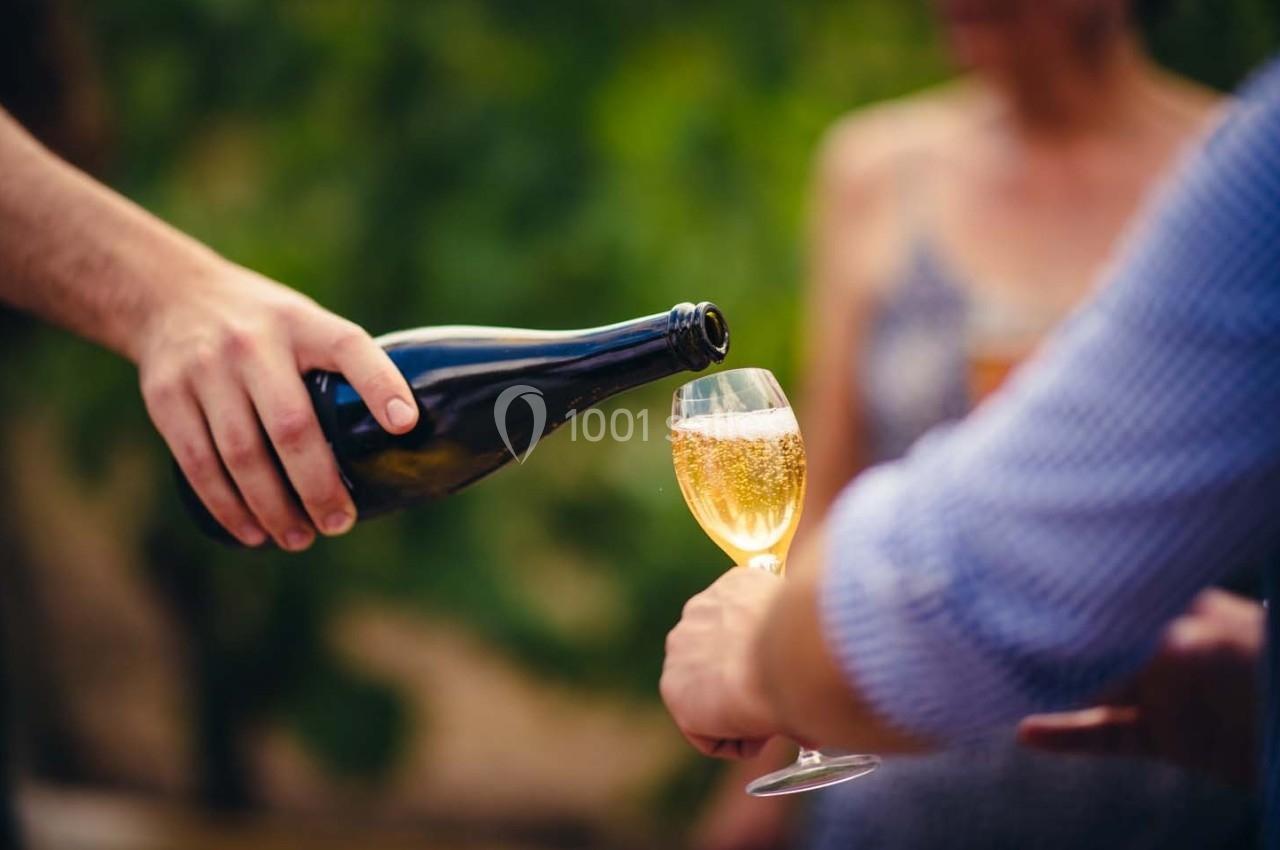 Une main verse du vin pétillant dans un verre tenu par une autre personne, en extérieur avec un arrière-plan flou.