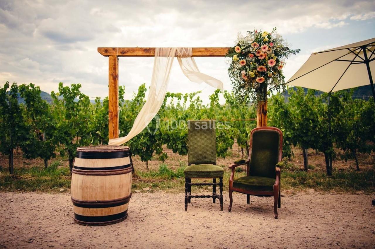 Deux chaises anciennes, un tonneau et une arche décorée de fleurs et de tissu, devant un vignoble.