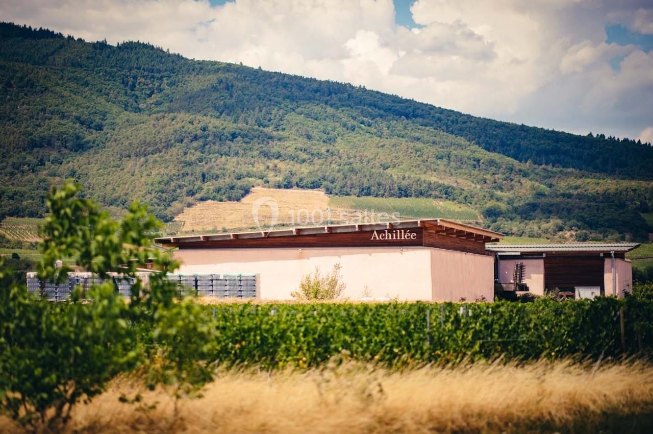 Bâtiment viticole entouré de vignes et de collines verdoyantes sous un ciel partiellement nuageux.