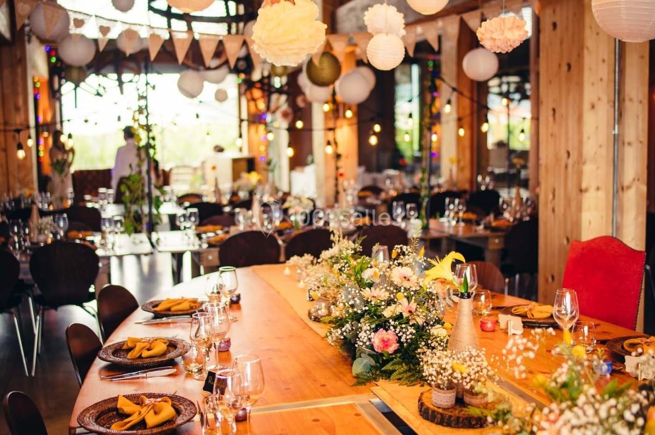 Salle décorée pour un événement, avec tables en bois ornées de fleurs, vaisselle et guirlandes lumineuses suspendues.