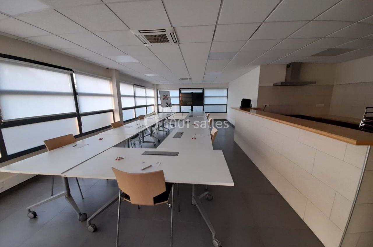 Salle de réunion lumineuse avec grandes tables blanches, chaises en bois clair et écran au fond.