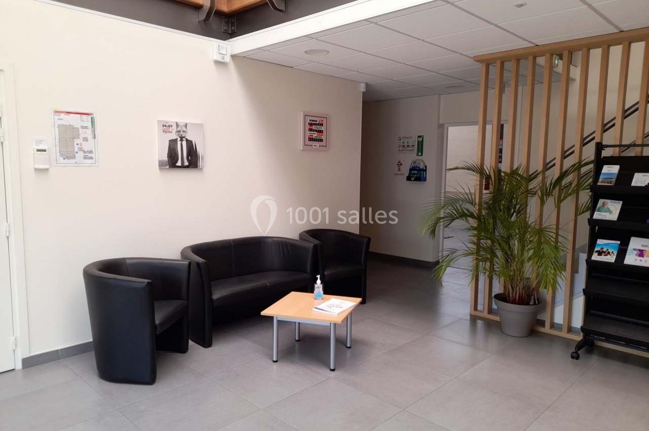 Espace d'attente avec fauteuils noirs, table basse, plante verte et affiches sur les murs dans un hall lumineux.