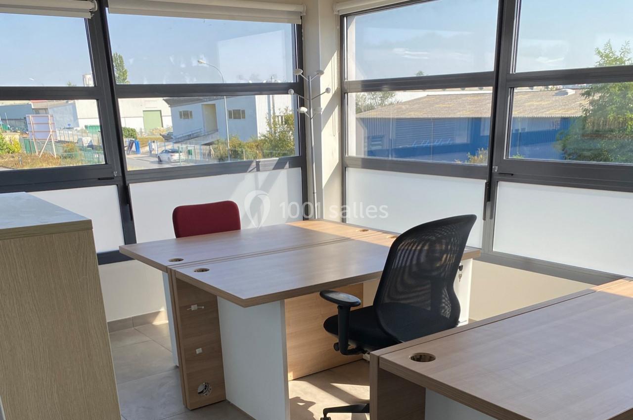 Bureau lumineux avec deux postes de travail, chaises et grandes fenêtres offrant une vue sur des bâtiments industriels.