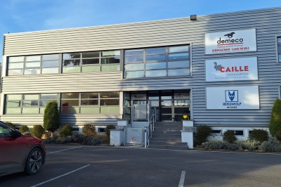 Bâtiment industriel moderne avec parking, panneaux d'entreprises visibles et végétation en premier plan.