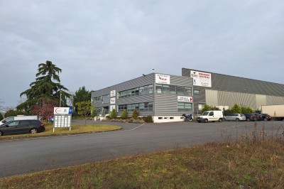 Bâtiment industriel moderne avec parking, panneaux d'entreprises visibles et végétation en premier plan.