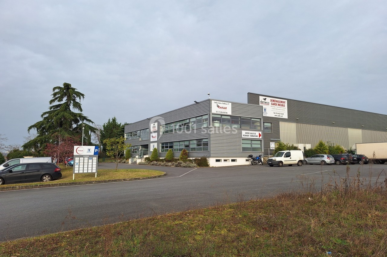 Bâtiment industriel moderne avec parking, panneaux d'entreprises visibles et végétation en premier plan.