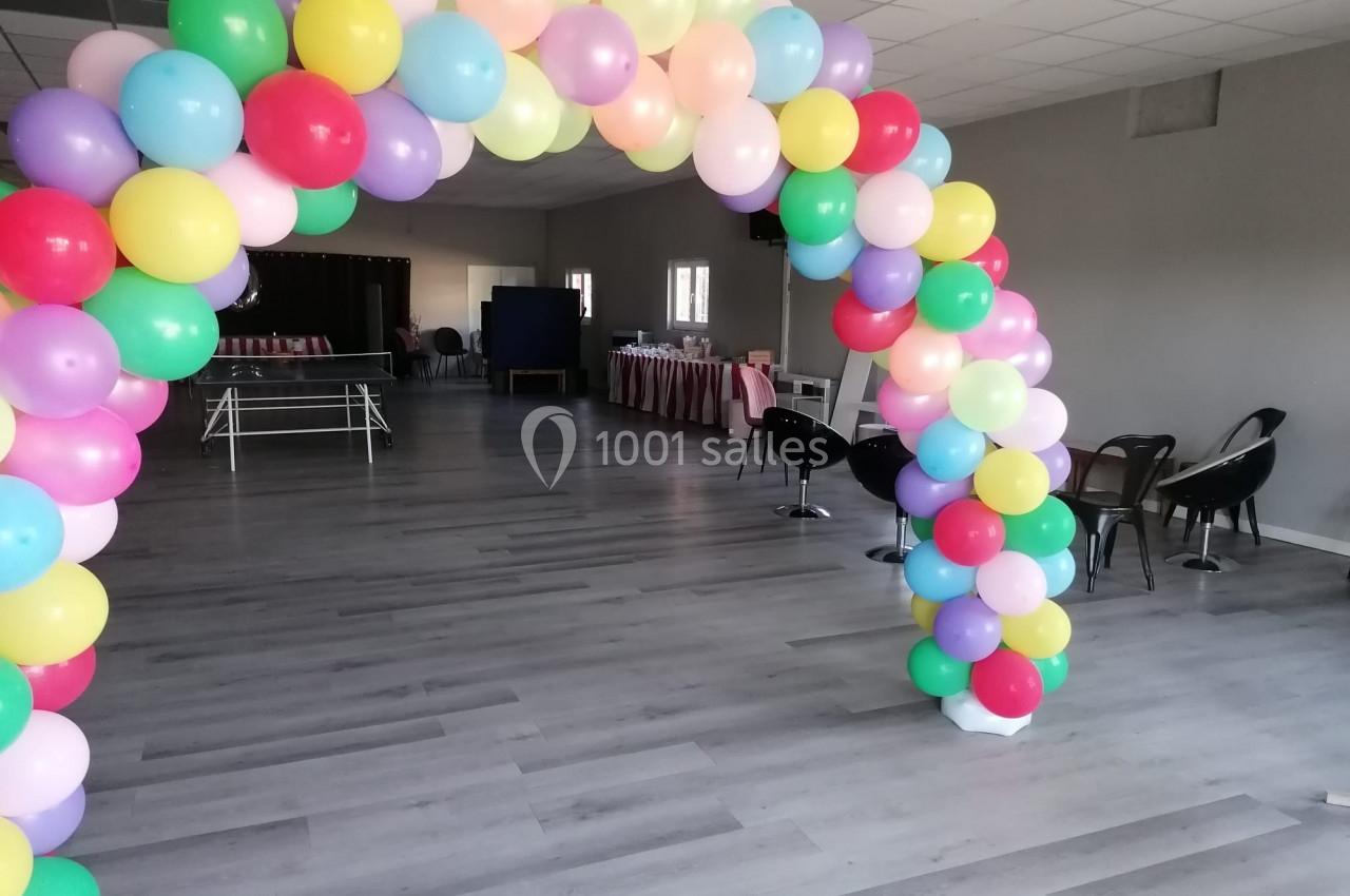 Arche de ballons multicolores dans une salle vide avec des tables et des chaises en arrière-plan.