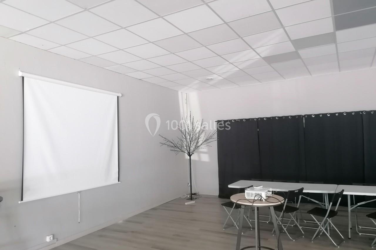 Salle lumineuse avec sol en bois, écran de projection, tables, chaises pliantes et rideaux noirs.