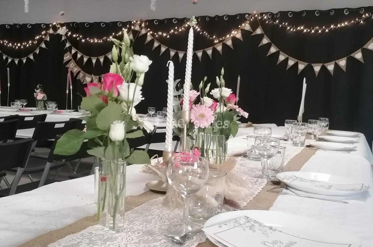 Salle décorée pour un événement avec tables dressées, nappes blanches, fleurs, bougies et guirlandes lumineuses.