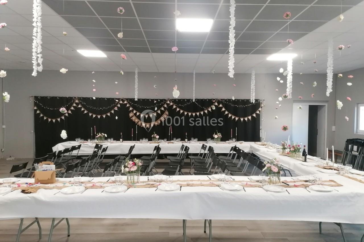 Salle décorée pour un événement, avec tables dressées, guirlandes suspendues et fleurs en centre de table.