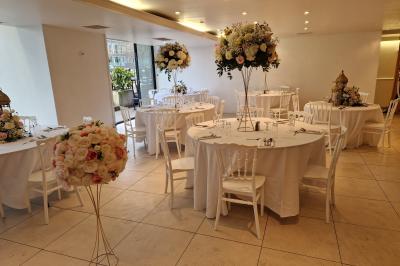 Centre de table floral avec roses et feuillage, entouré de tables dressées dans une salle lumineuse.