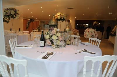 Centre de table floral avec roses et feuillage, entouré de tables dressées dans une salle lumineuse.