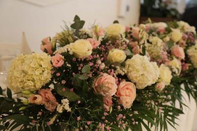Centre de table floral avec roses et feuillage, entouré de tables dressées dans une salle lumineuse.