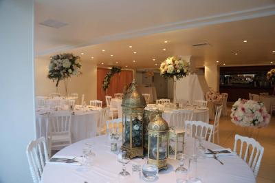 Centre de table floral avec roses et feuillage, entouré de tables dressées dans une salle lumineuse.