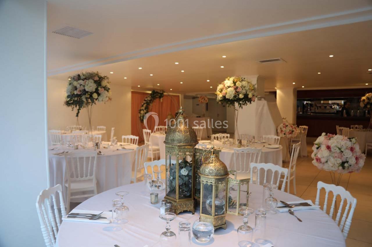 Salle de réception décorée avec des tables rondes, nappes blanches, lanternes dorées et compositions florales.