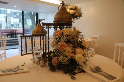 Centre de table floral avec roses et feuillage, entouré de tables dressées dans une salle lumineuse.