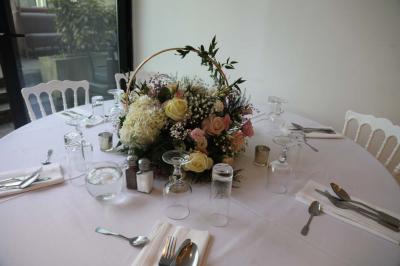 Centre de table floral avec roses et feuillage, entouré de tables dressées dans une salle lumineuse.