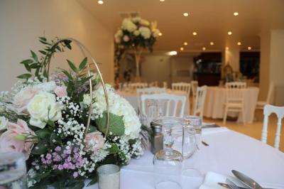 Centre de table floral avec roses et feuillage, entouré de tables dressées dans une salle lumineuse.