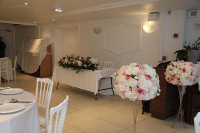 Centre de table floral avec roses et feuillage, entouré de tables dressées dans une salle lumineuse.