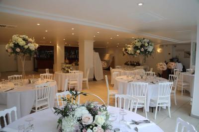 Centre de table floral avec roses et feuillage, entouré de tables dressées dans une salle lumineuse.
