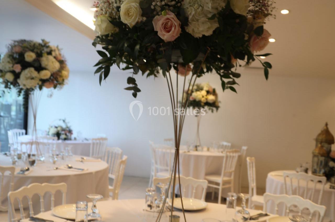 Centre de table floral avec roses et feuillage, entouré de tables dressées dans une salle lumineuse.