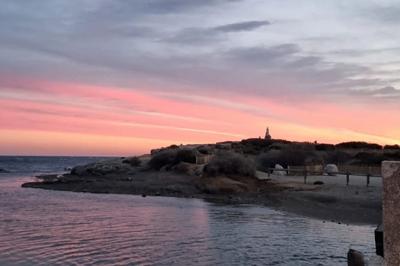 Vue d'une plage avec une terrasse ombragée, des promeneurs, une étendue d'eau et des collines à l'horizon.