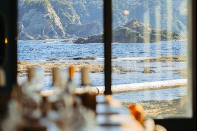 Vue d'une plage avec une terrasse ombragée, des promeneurs, une étendue d'eau et des collines à l'horizon.