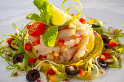 Vue d'une plage avec une terrasse ombragée, des promeneurs, une étendue d'eau et des collines à l'horizon.