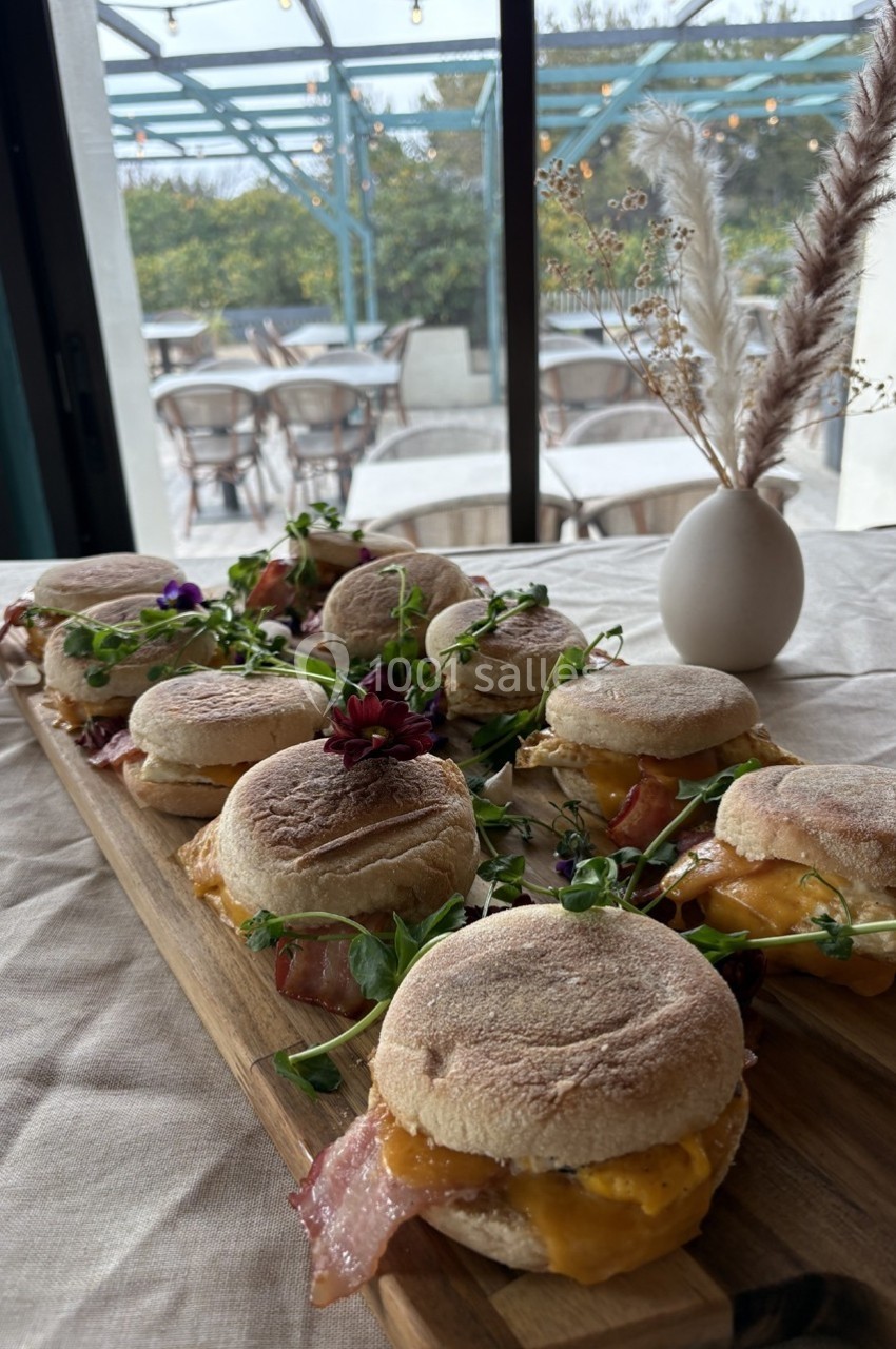 Plateau de petits sandwichs garnis de bacon, fromage et herbes fraîches, posé sur une table près d'une baie vitrée.