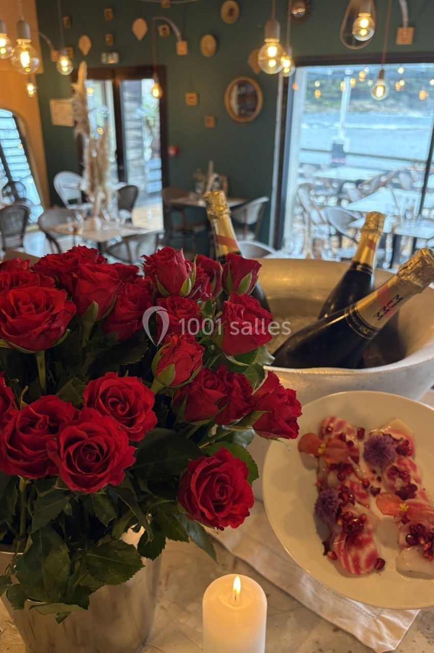Bouquet de roses rouges près d'une bougie allumée, avec un plat raffiné et des bouteilles de champagne en arrière-plan.