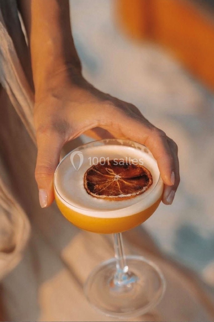 Main tenant un cocktail jaune garni d'une tranche d'orange séchée, en extérieur avec une lumière douce.