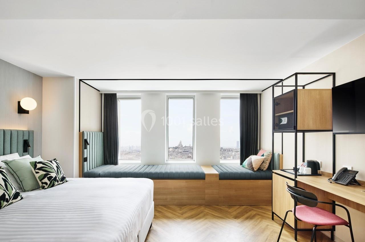 Chambre moderne avec lit double, coin bureau, banquette près des fenêtres et vue sur la ville.