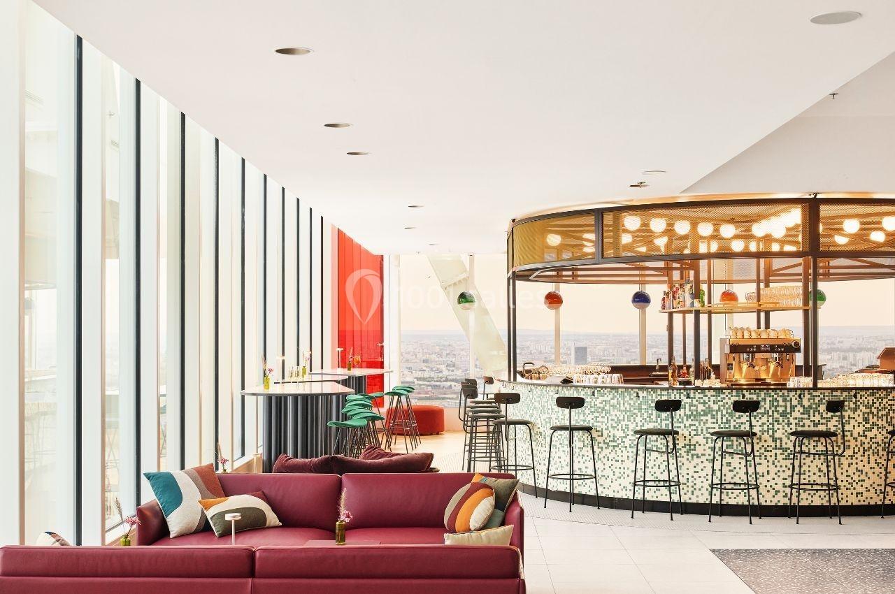 Espace lumineux avec un bar circulaire moderne, des tabourets et un canapé rouge, vue sur la ville en arrière-plan.