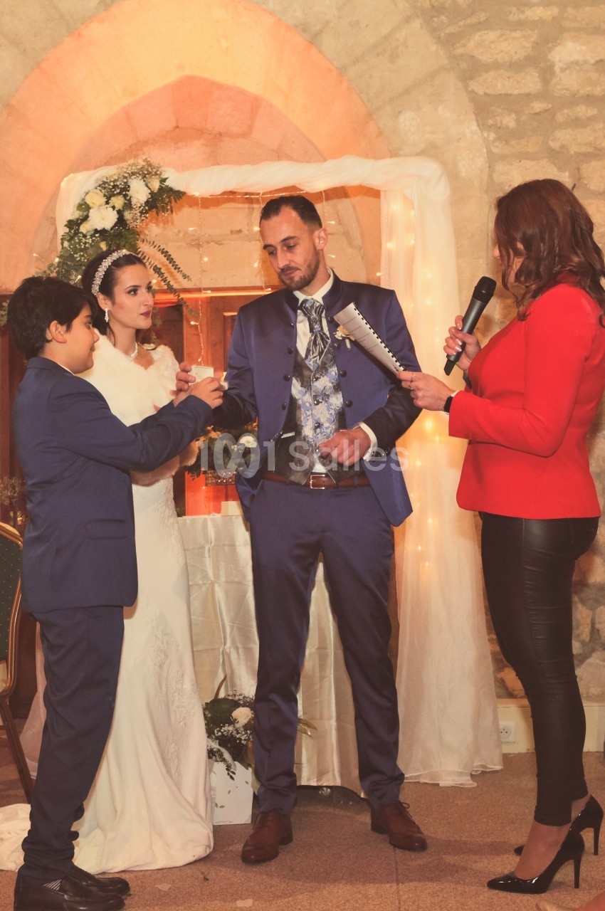 Un couple en tenue de mariage échange des vœux devant une femme avec un micro et un jeune garçon dans une salle en pierre.
