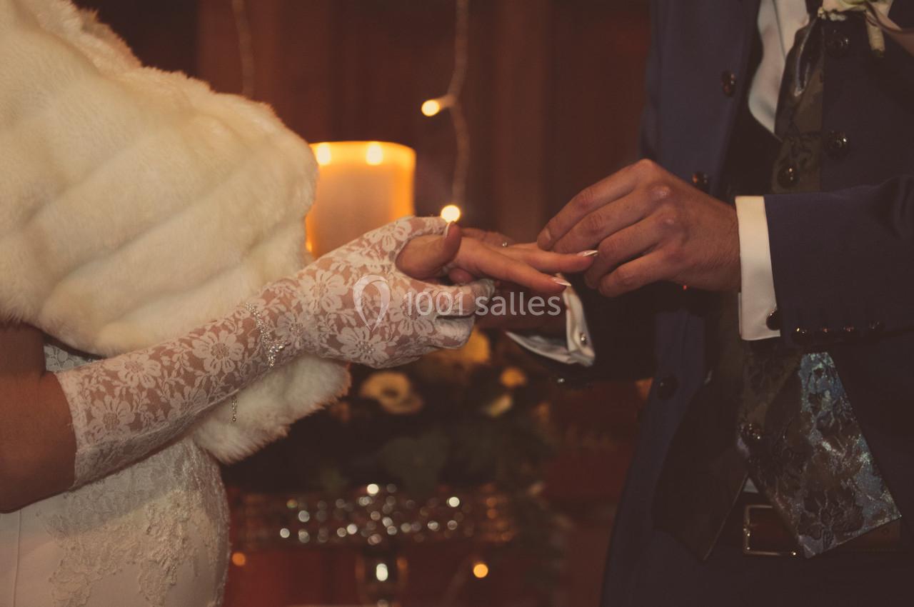 Un marié passe une alliance au doigt de la mariée portant des gants en dentelle, devant une bougie allumée.