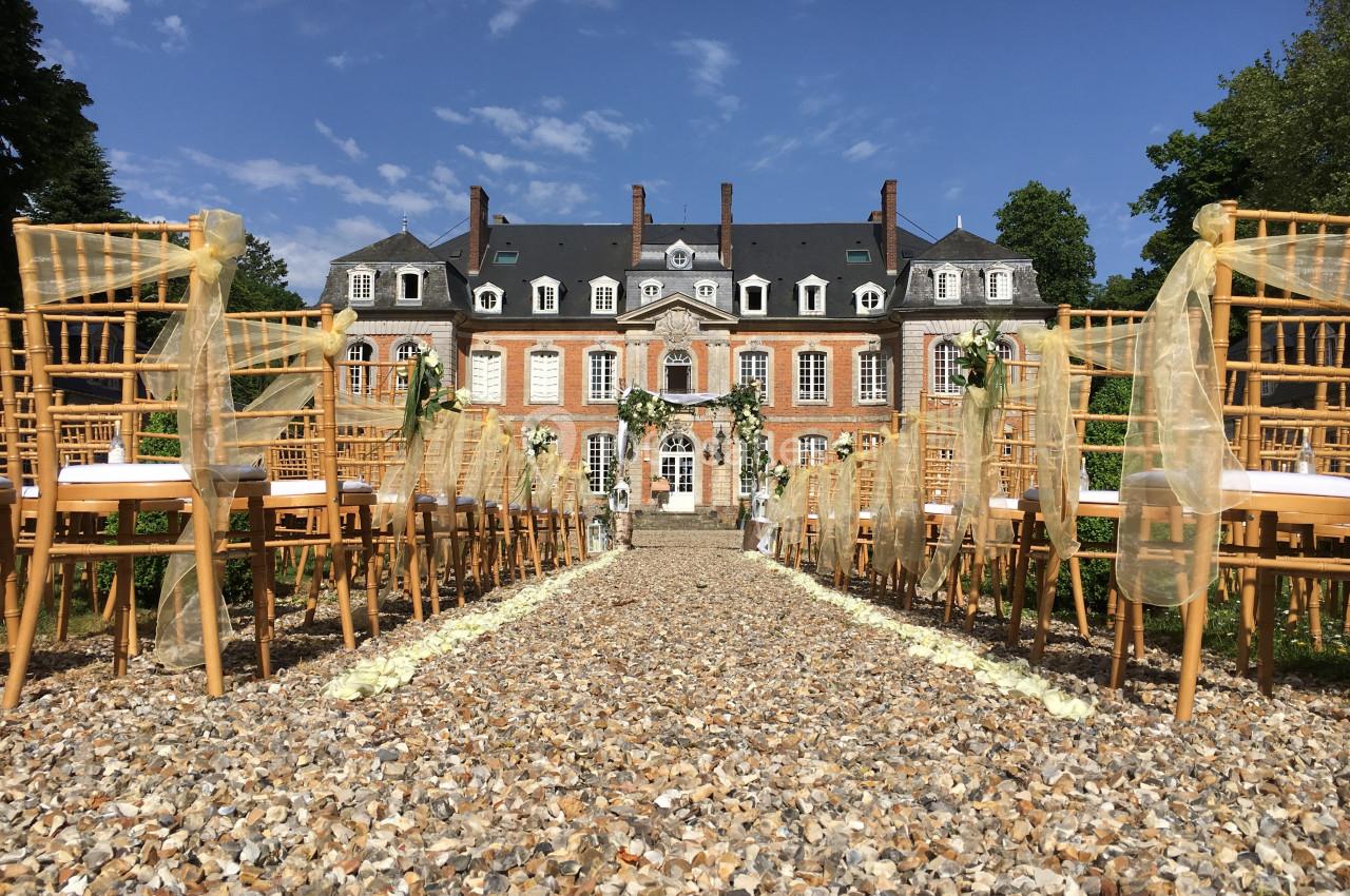Allée centrale décorée pour une cérémonie en plein air devant un château en briques rouges par temps ensoleillé.