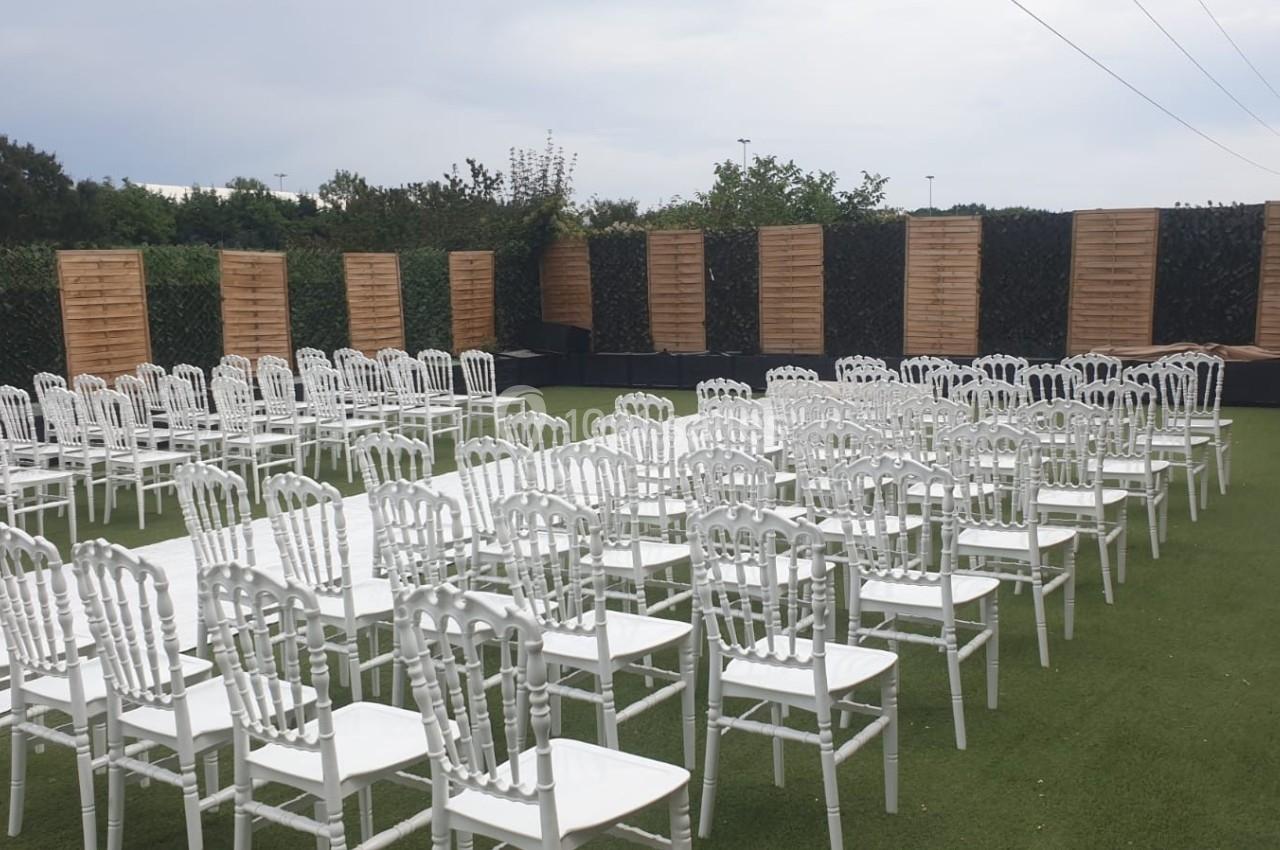 Chaises blanches alignées sur une pelouse artificielle, disposées face à une scène extérieure entourée de panneaux en bois.