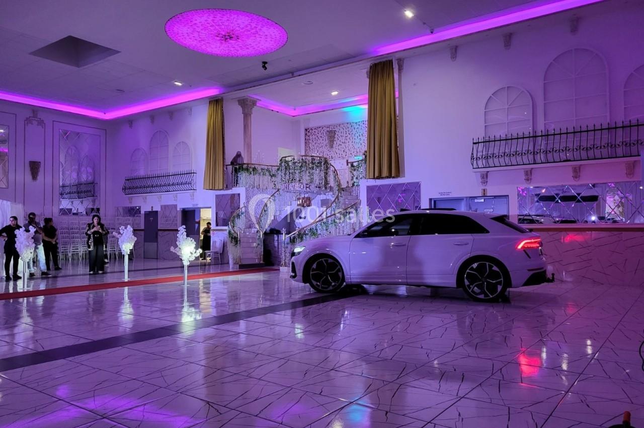 Voiture blanche garée dans une grande salle de réception éclairée par des lumières violettes avec décorations élégantes.