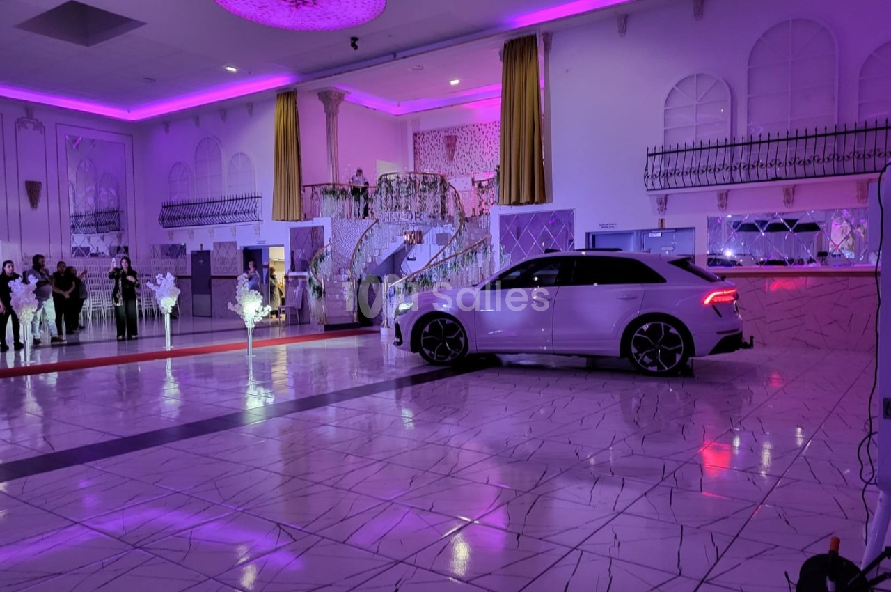 Voiture blanche garée dans une grande salle de réception illuminée par des lumières violettes, avec des invités en arrière…