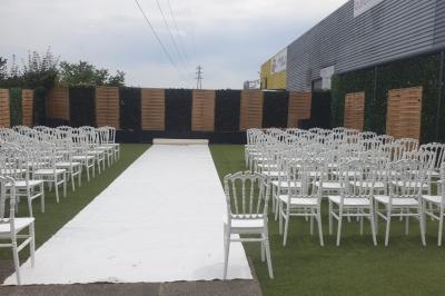 Allée centrale bordée de chaises blanches disposées en extérieur, entourée de panneaux en bois et de murs végétalisés.