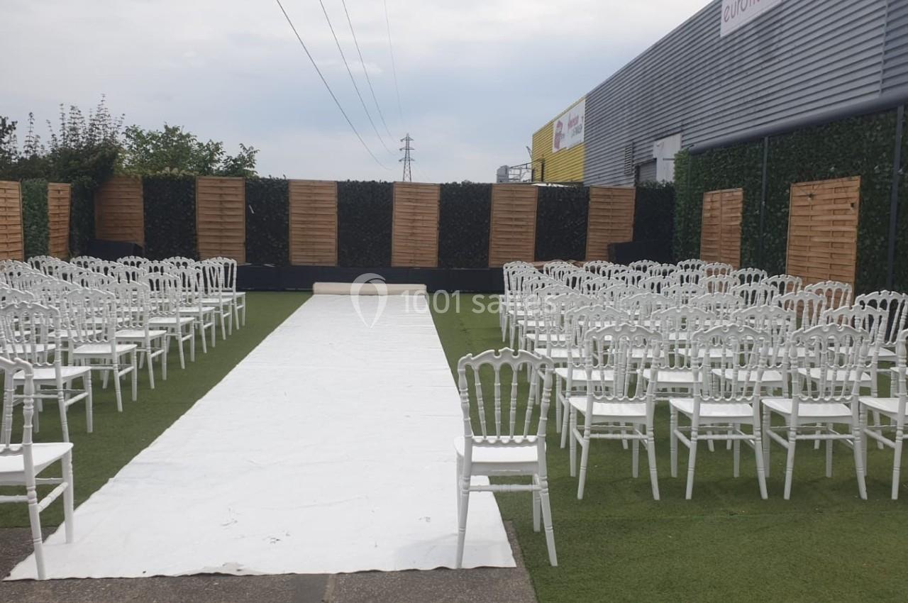 Allée centrale bordée de chaises blanches disposées en extérieur, entourée de panneaux en bois et de murs végétalisés.