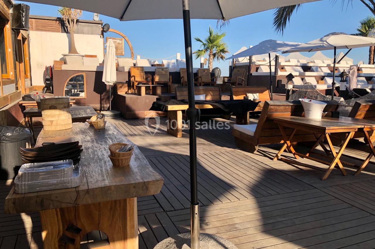 Terrasse en bois avec tables et chaises, parasols ouverts, et vue sur des palmiers sous un ciel dégagé.