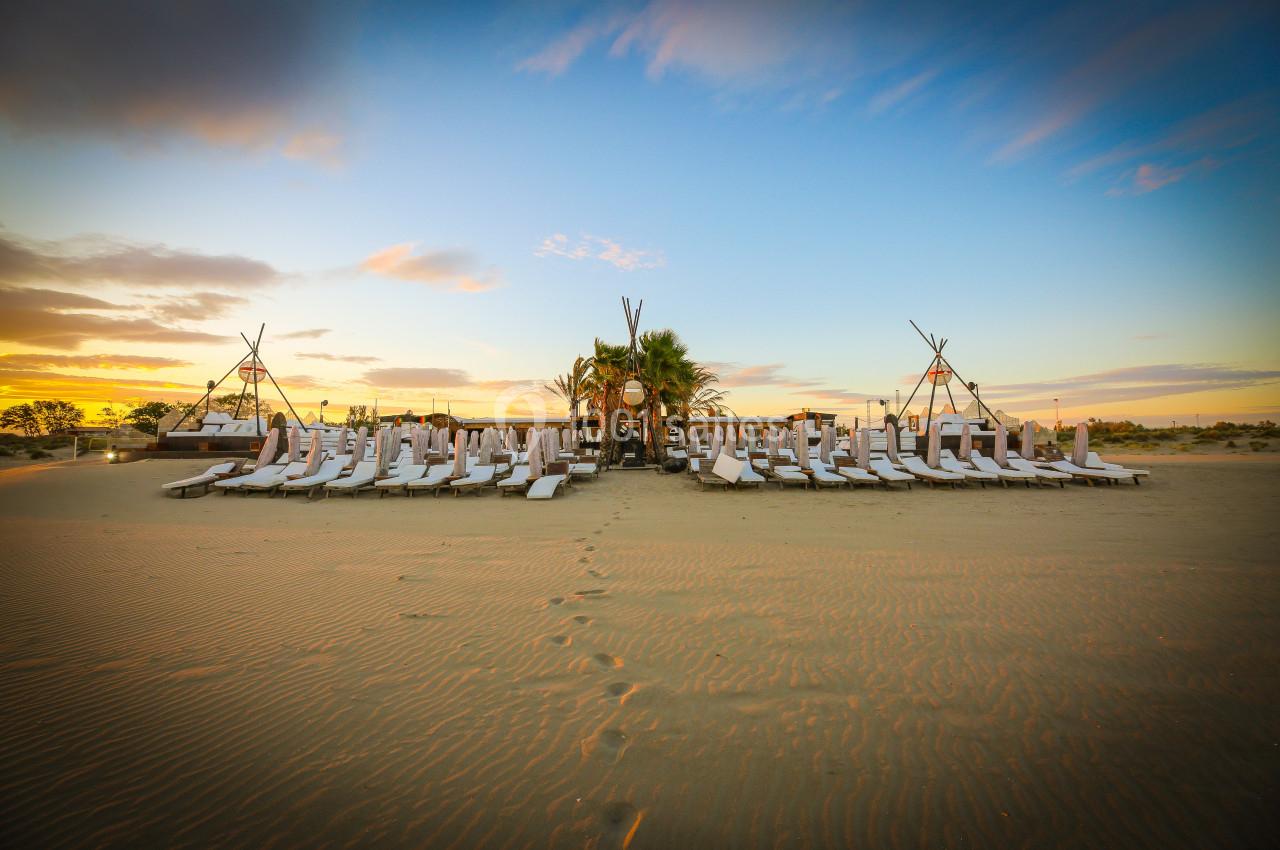 Alignement de transats blancs sur une plage de sable au coucher du soleil, avec des structures en bois en arrière-plan.