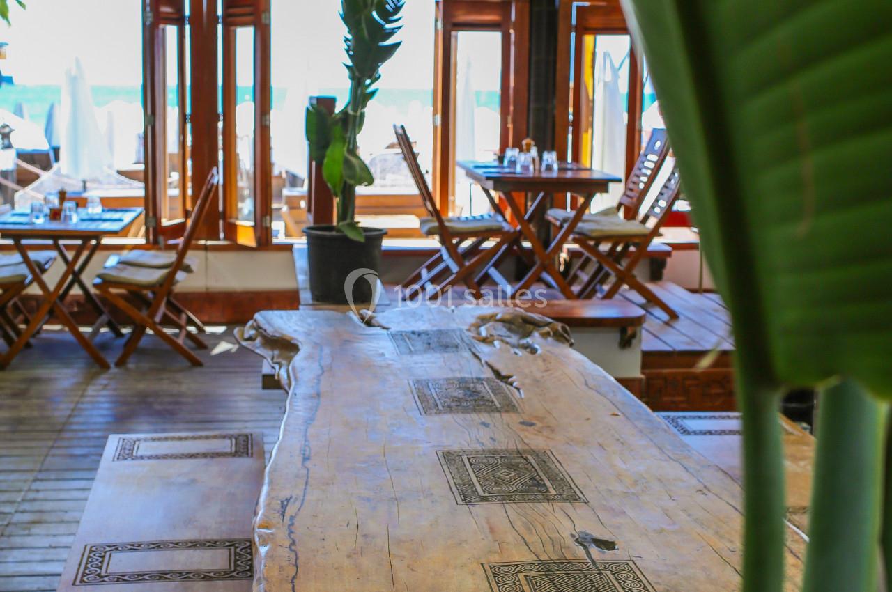 Grande table en bois brut ornée de motifs gravés, dans un espace lumineux avec des chaises pliantes et des plantes.