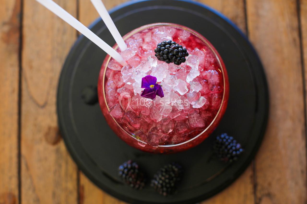 Verre de cocktail rouge avec glace pilée, mûres fraîches et une fleur violette, posé sur une table en bois.